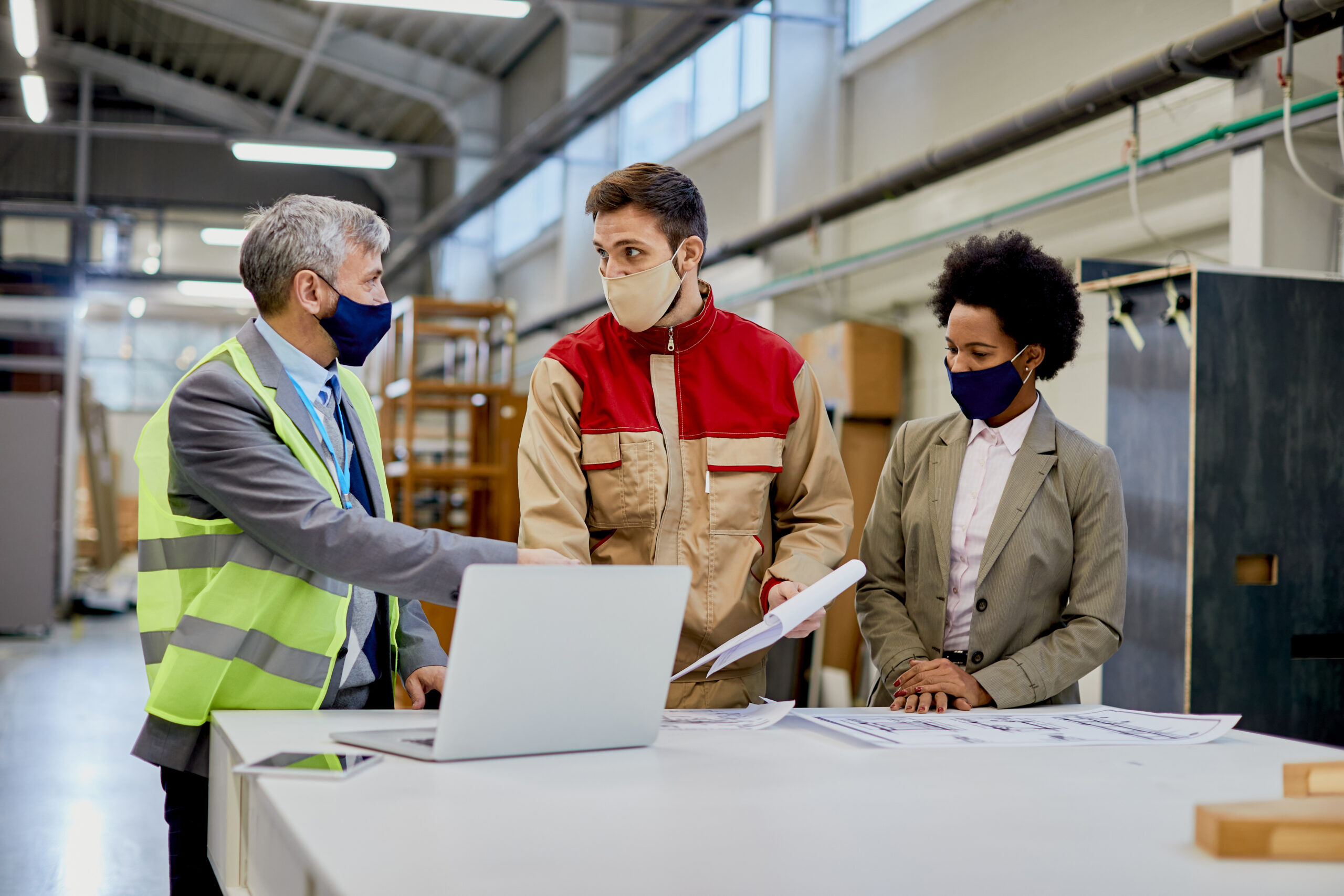 Woodworking factory laborer and quality control inspectors wearing protective face masks while talking and analyzing paperwork at production facility.