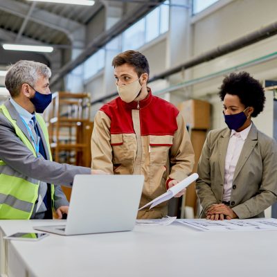 Woodworking factory laborer and quality control inspectors wearing protective face masks while talking and analyzing paperwork at production facility.